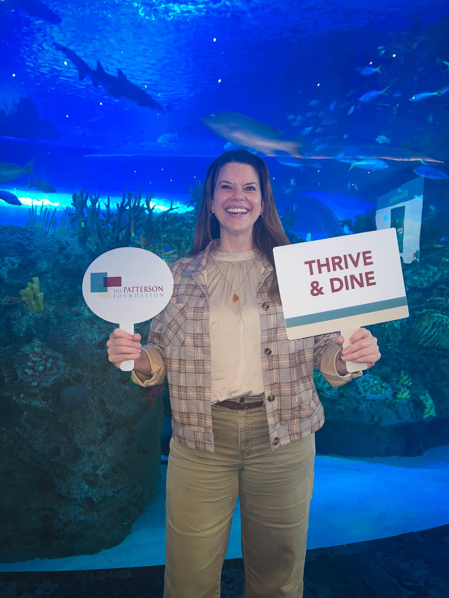 Executive Director Annie Berkness holding sign with logo of Patterson Foundation on left and sign reading, "Thrive and Dine," on right.