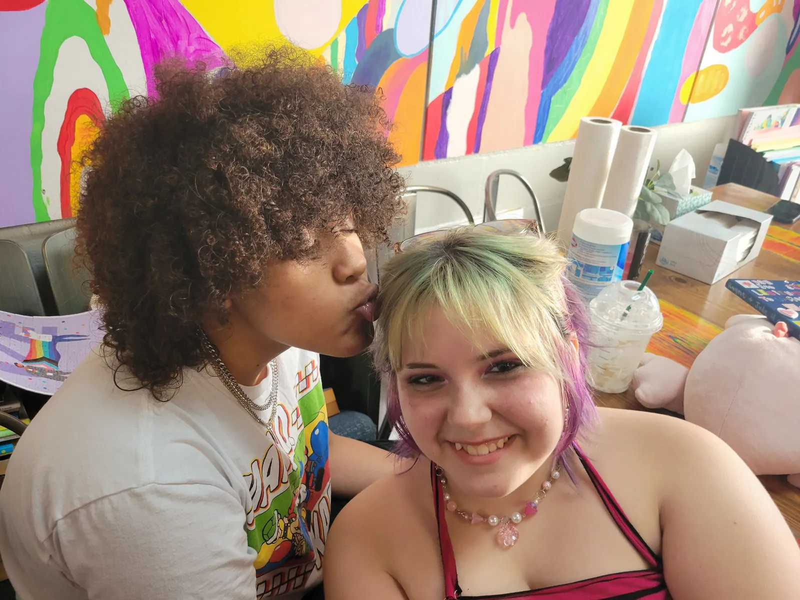 Two friends in front of a colorful mural, one kissing the top of the other's head.