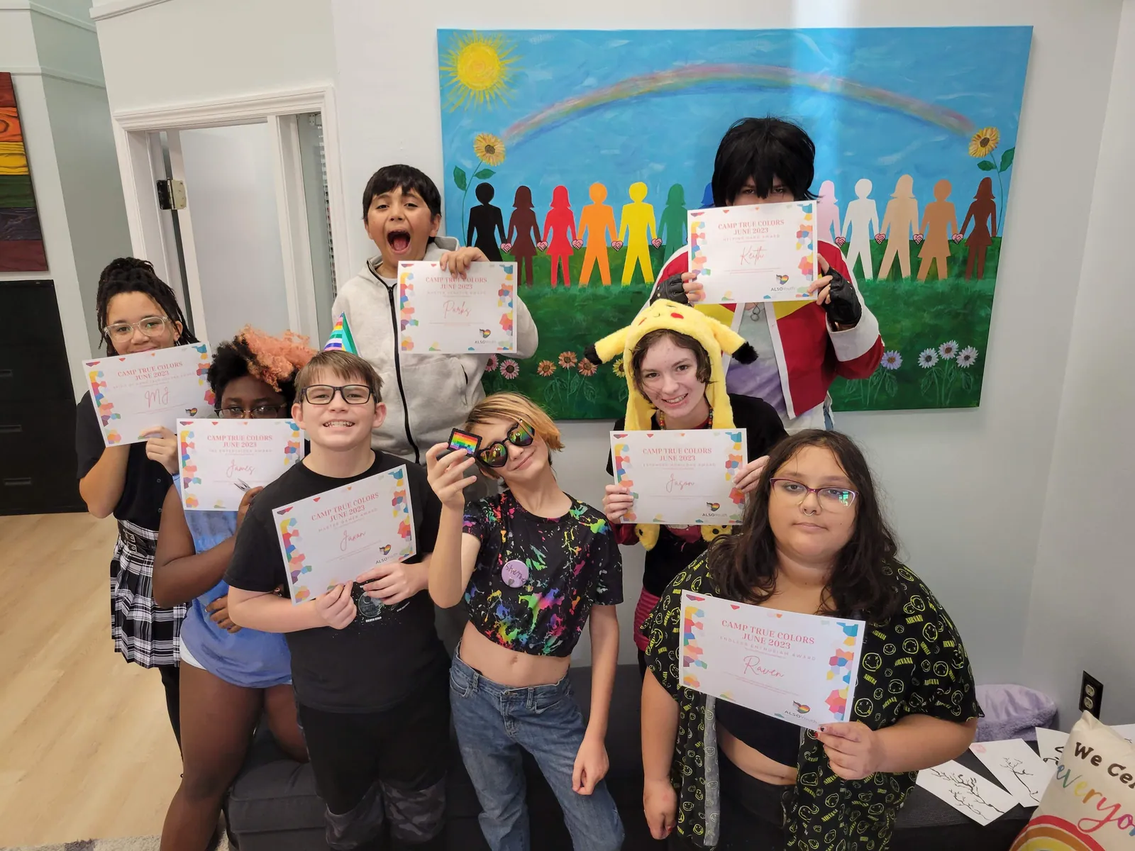 Campers holding up their certificates in front of a rainbow mural of people holding hands.