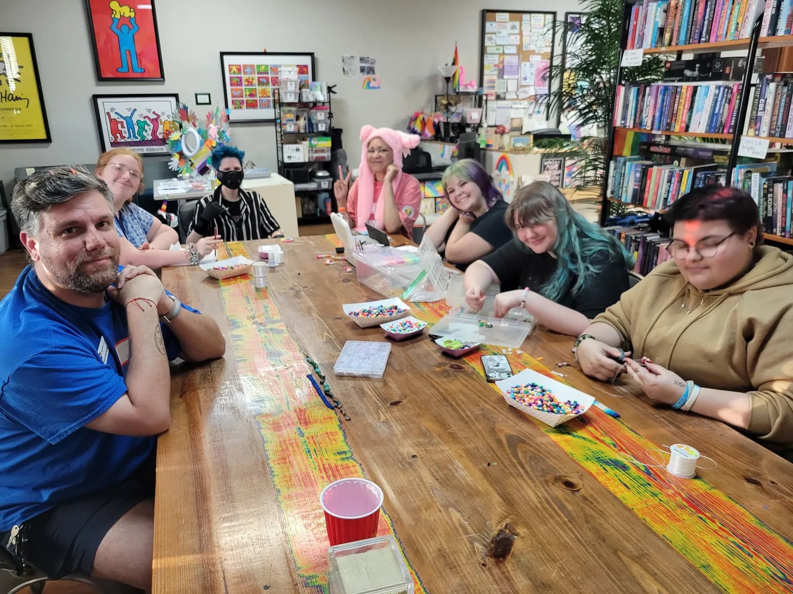 Young people and an adult around a long painted table with snacks and crafts, in a room lined with books and art.