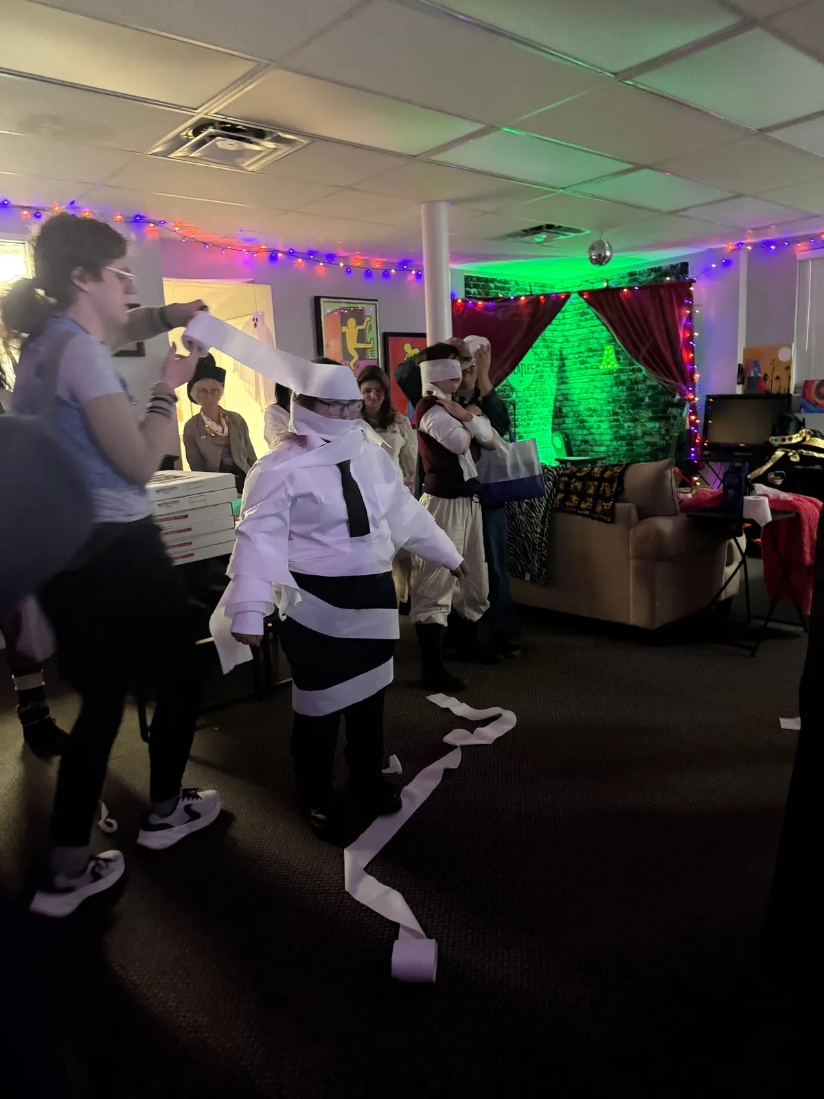 Costumed guests mid game at Halloween, one mummy wrapped in paper and a black tie, under purple lights and a green lit stage.