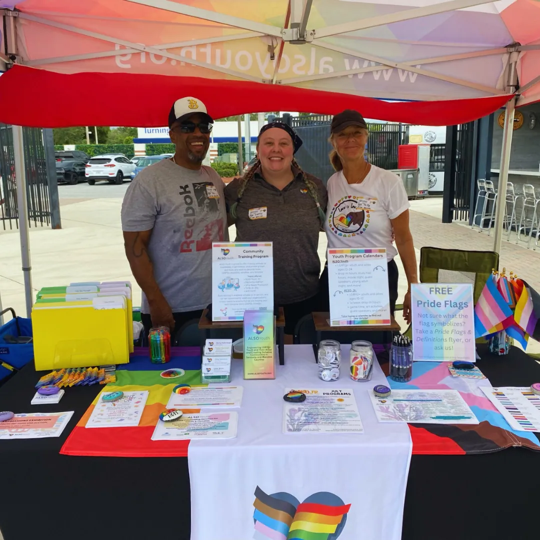 Three volunteers at the ALSO Youth outreach table, covered in Pride flags and flyers.