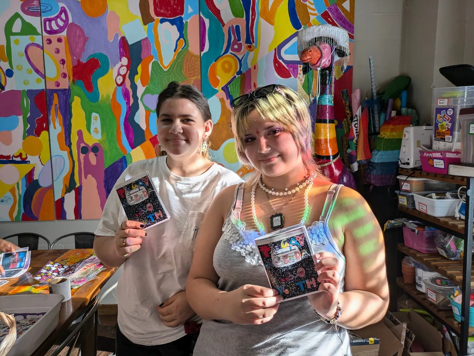 Two young people proudly holding up the zines they made, in front of a wildly colorful painted wall.