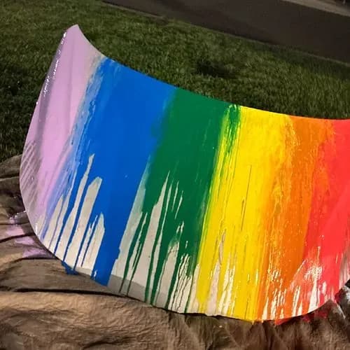 A car hood lying on a drop cloth in the grass, painted with dripping stripes of pink, blue, green, yellow, orange, and red.