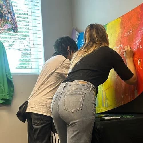 Two people, seen from behind, writing on the rainbow painted car hood where it hangs on a wall. Words like Hope and Love are already written on it.