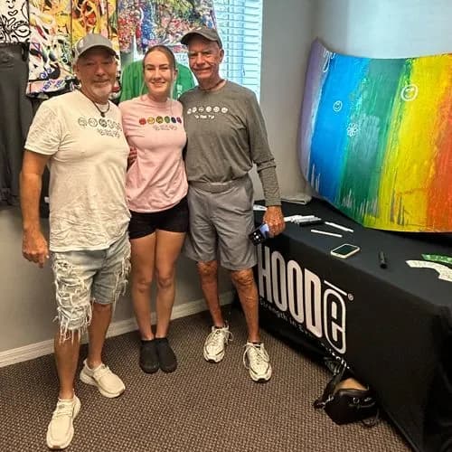Three people smiling in a room next to the painted car hood and a table with a HOODē banner and markers.