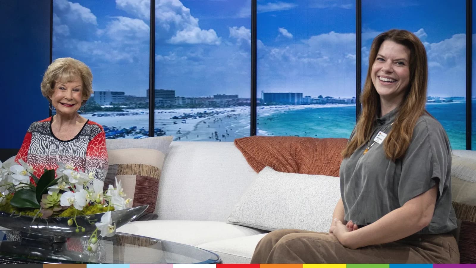 Two women smiling on a bright TV studio set, with a sunny beach view on the screens behind them.