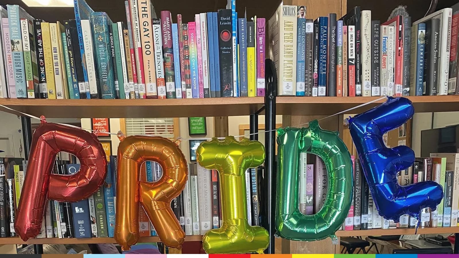 Rainbow foil balloons spelling PRIDE hang across shelves full of books at an ALSO Youth center.