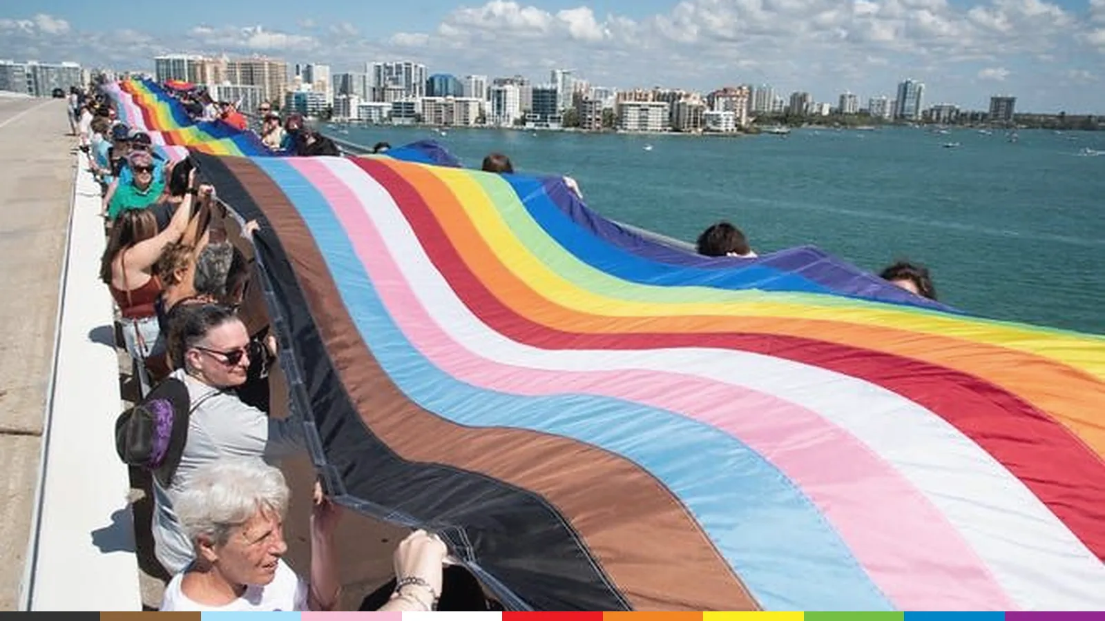 People carry a giant Progress Pride flag along the Sarasota waterfront, with the downtown skyline across the bay.