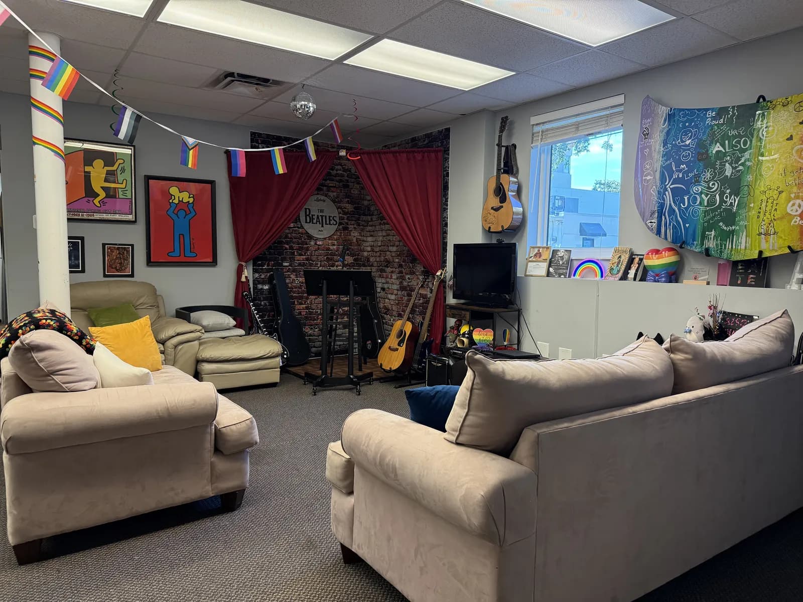 A cozy corner of the Sarasota Center lounge with couches, red curtains, a brick wall, guitars, and pride flags overhead.