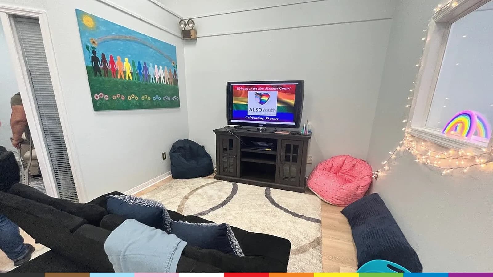 The youth lounge at ALSO Youth's Manatee Center: couches, bean bags, string lights, and a mural of people holding hands under a rainbow.