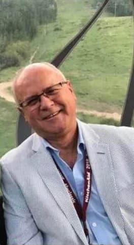 Greg Jung smiling outdoors in a light blazer, with a green mountainside behind him.